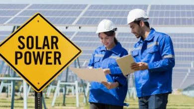 Two engineers in blue uniforms and white helmets working at a solar power plant, reviewing documents with solar panels in the background and a yellow "Solar Power" sign in the foreground.