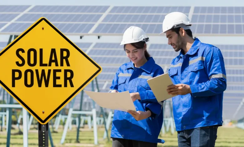 Two engineers in blue uniforms and white helmets working at a solar power plant, reviewing documents with solar panels in the background and a yellow "Solar Power" sign in the foreground.