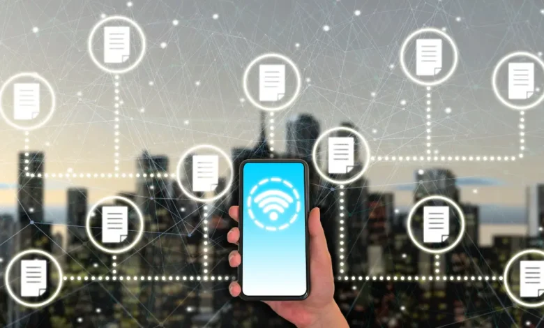 A hand holding a smartphone with a wireless icon on screen, surrounded by floating document icons symbolizing data transfer in a digital network over a city background.
