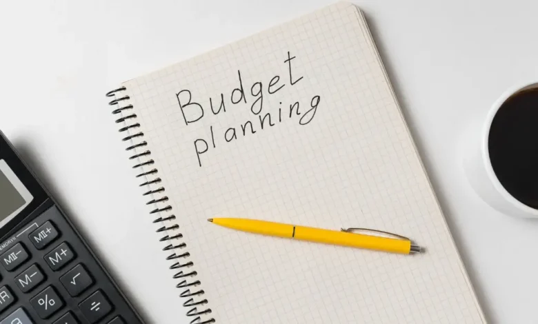 Notebook with "Budget planning" written on it, placed beside a yellow pen, calculator, and a cup of coffee on a white desk.